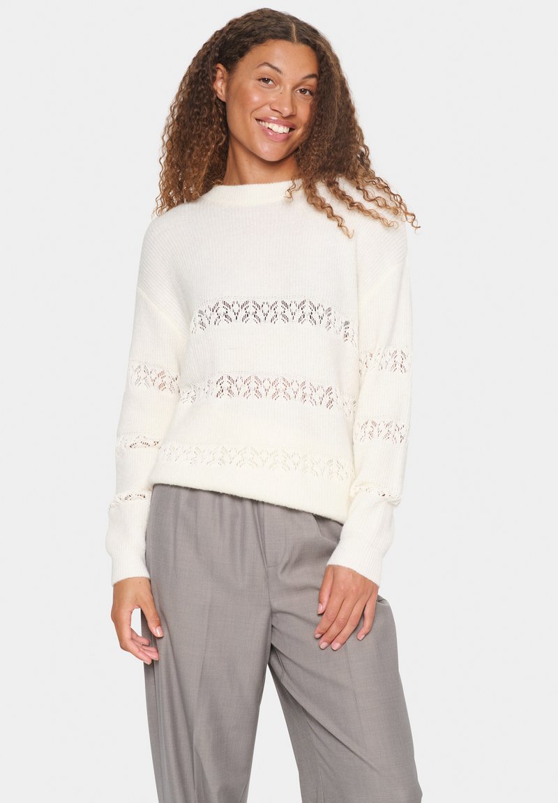 White knitted sweater featuring horizontal lace patterns, round neckline, and long sleeves, paired with light grey trousers.