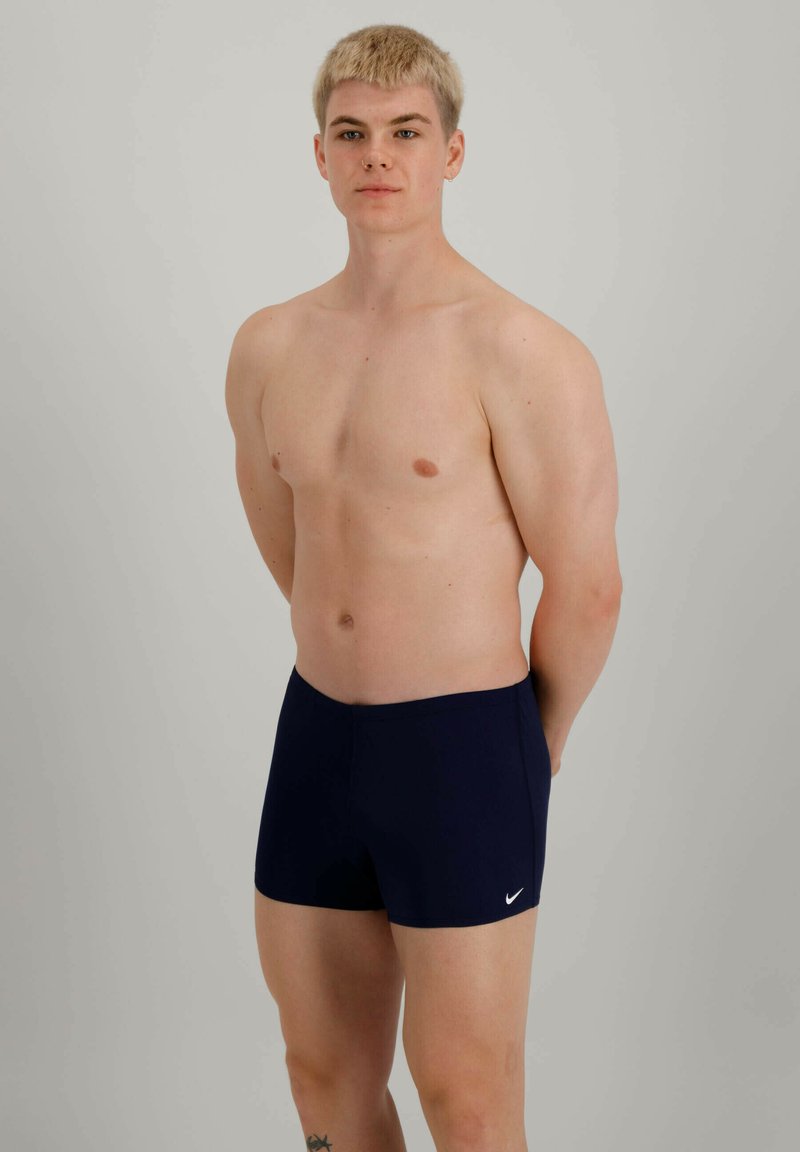 Sötétkék fürdősort elasztikus anyagból, testhez álló fazonnal, fehér Nike logóval a bal elülső részen.
