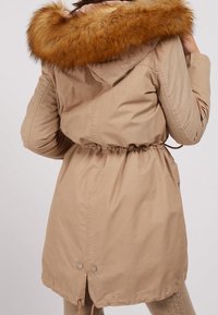 Kvinde iført beige vinterparka med stor brun pelskantet hætte, indsnøret talje og med manchetter på ærmerne, vist bagfra.