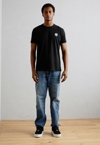 T-shirt en coton noir avec un petit emblème, associé à un jean en denim bleu clair. Le modèle porte des baskets noires avec des semelles blanches.