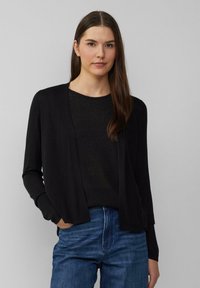 Zwarte open voorzijde cardigan van zacht materiaal, met lange mouwen en een ontspannen, iets oversized pasvorm, gecombineerd met een donkere trui.