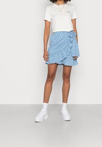 T-shirt blanc avec texte coloré, associé à une jupe bleue à volants ornée d'un motif floral blanc. Porté avec des baskets blanches et des chaussettes.