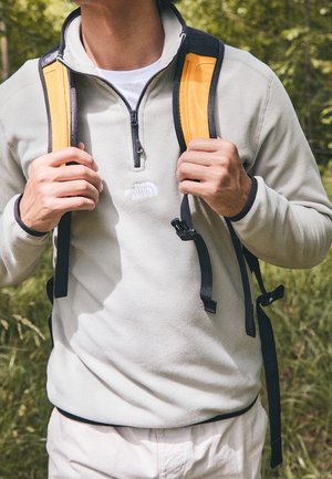 Vaaleanharmaa fleece-puolipaita, jossa on puolivetoketju ja rintamuksessa logo. Edessä pidetään mustaa ja oranssia reppuhihnaa.