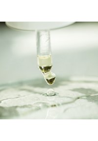 Pipette, die eine goldene Flüssigkeit auf eine klare, reflektierende Oberfläche träufelt. Der Tropfen bildet beim Aufprall einen kleinen Tropfen. Weichgezeichneter Hintergrund.