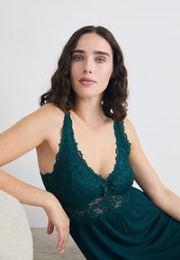 Nainen, jolla on kihara tumma hius, pukeutuneena teal-väriseen pitsitoppiin, istuu ja nojaa vaalean tekstuurin omaavaan pintaan yksinkertaista taustaa vasten.