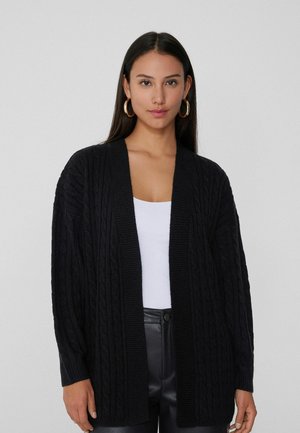 Cardigan en tricot noir à mailles torsadées, avec une ouverture à l'avant, des bords côtes et des manches longues. Porté sur un haut blanc et associé à un pantalon en cuir noir.