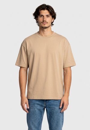 Mężczyzna o krótkich ciemnych włosach, ubrany w jednolity beżowy, oversize'owy t-shirt i niebieskie dżinsy, stojący na jasnoszarym tle.