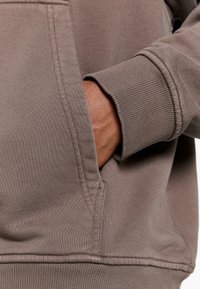 Brown hoodie with ribbed cuffs and waistband, featuring a side pocket. Material has a soft, textured appearance.