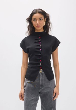 Mujer con cabello oscuro y ondulado lleva una camiseta negra de manga corta con botones multicolores y vaqueros grises con tachuelas doradas en el bolsillo.