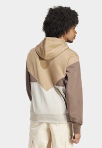 Man med lockigt hår som bär en mångtonad hoodie i beige, brunt och vitt, stående på sidan med händerna avslappnat vid sidorna.