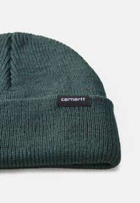 Căciulă din tricot ribbed de culoare verde închis, dotată cu un tiv pliabil, pe care se află un eticheta neagră cu brandul "carhartt."