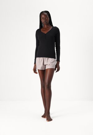 Svart långärmad henley-topp med tre knappar, kombinerad med ljusrandiga shorts. Modellen står barfota mot en enfärgad vit bakgrund.