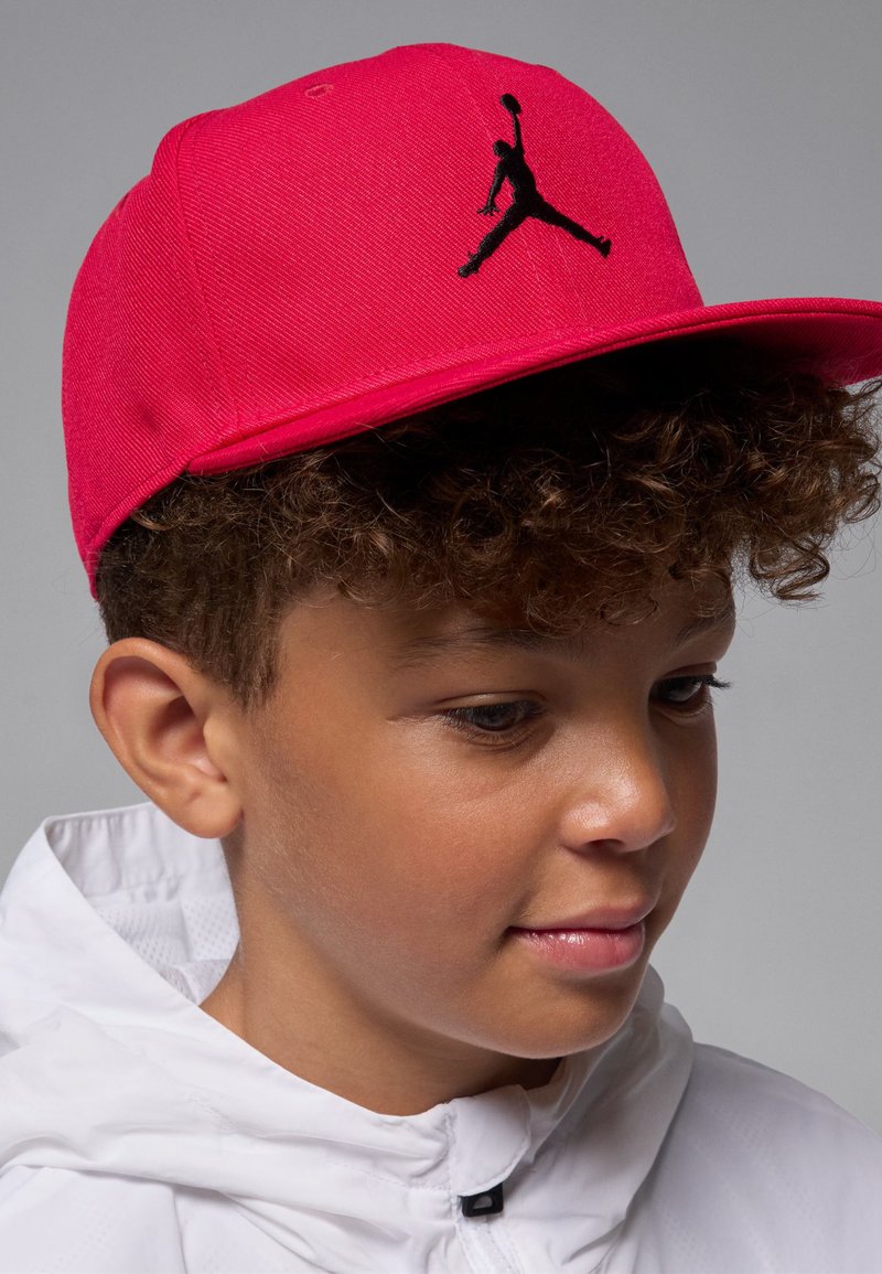 Red baseball cap with a black Jumpman logo on the front; features a structured brim and fabric texture, worn by a child in a white jacket.