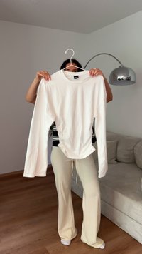 Bela dolgosleeve majica s okroglim izrezom, z nabranimi detajli na strani in gladkim, lahkim materialom, viseča na obešalniku.