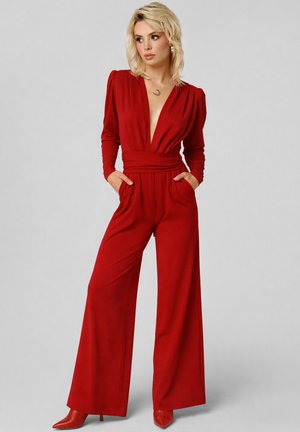 Donna bionda con le mani nelle tasche, indossa una tuta rossa a maniche lunghe con scollo a V profondo, pantaloni ampi e tacchi rossi abbinati.