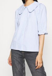 Blouse rayée bleu et blanc avec un grand col et des manches courtes bouffantes ; elle possède une fermeture à boutons sur le devant et une coupe décontractée.