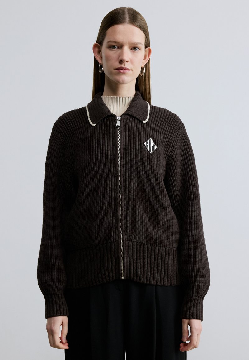 Pull en tricot côtelé marron avec fermeture éclair, col blanc contrastant et logo en forme de losange sur le côté gauche de la poitrine. Texture douce.