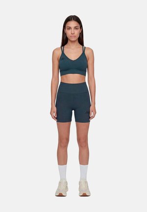 Femme debout face à l'avant, portant un soutien-gorge de sport bleu sarcelle foncé et un short cycliste taille haute avec des chaussettes blanches et des baskets beiges sur fond blanc.