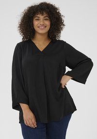 Black, loose-fitting blouse with a V-neck, wide sleeves, and a pocket on one side. Fabric has a soft texture, paired with blue jeans.