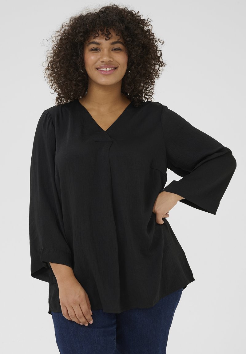 Black, loose-fitting blouse with a V-neck, wide sleeves, and a pocket on one side. Fabric has a soft texture, paired with blue jeans.