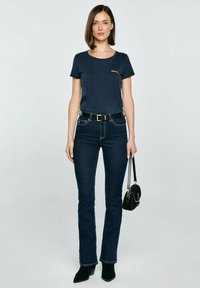 Námořnická modrá tričko s kulatým výstřihem, doplněné tmavými džíny s volnými nohavicemi. Černý opasek se zlatou sponou, držící malou černou kabelku.