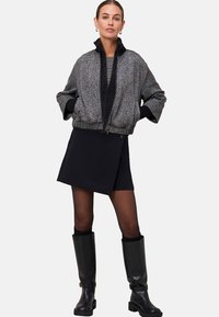 Femme portant une veste zippée à motifs, une jupe portefeuille noire, des collants transparents et des bottes noires à hauteur de genoux, se tenant avec les mains dans les poches de la veste.