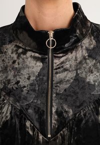 Black floral patterned zip-up top with a high collar, featuring a gold zipper and textured fabric. Ruffled detail at the hem.