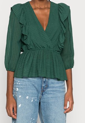 Grøn bluse med dyb V-udskæring, flæse detaljer langs skuldrene og foran, med en tekstureret stof og peplum talje, kombineret med blå jeans.