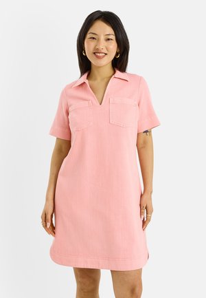 Femme souriante portant une robe rose à manches courtes avec col et deux poches poitrine, debout devant un fond clair uni.