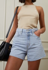 Top sin mangas de canalé beige combinado con pantalones cortos de denim azul claro, con cintura alta, bolsillos frontales y cierre de botón. Bolso negro con detalles dorados.