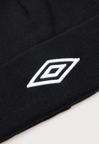 Melnas adītas audums ar baltu izšūtu dubultā dimanta logotipu tuvu apakšējai malai, uz vienkārša gaiša fona.