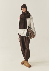 Witte gebreide cardigan met bloemenborduursel, bruine oversized broek, bijpassende sjaal en hoed, en bruine enkellaarsjes. Getoonde tan handtas.