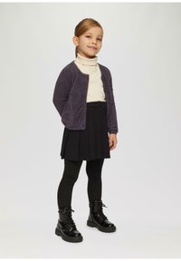 Kind, der einen lila flauschigen Cardigan, einen cremefarbenen Rollkragenpullover, einen schwarzen Faltenrock und schwarze Schnürstiefel mit klobigen Sohlen trägt.