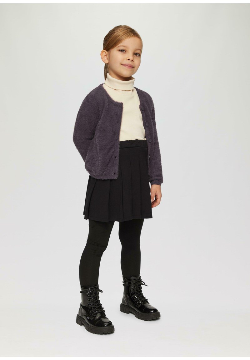 Kind, der einen lila flauschigen Cardigan, einen cremefarbenen Rollkragenpullover, einen schwarzen Faltenrock und schwarze Schnürstiefel mit klobigen Sohlen trägt.
