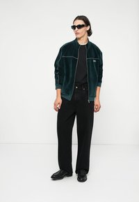 Jonge man in een donkergroene fluwelen zip-up jack, zwart T-shirt, brede zwarte jeans, zwarte schoenen en zwarte zonnebril, staande tegen een witte achtergrond.