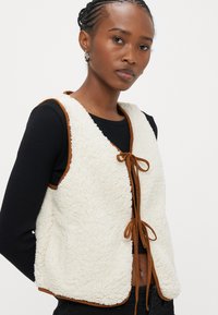 Fuskepelsvest i krem med brune kanter, med V-hals, knyting foran og myk tekstur. Brukt over en svart langermet topp.