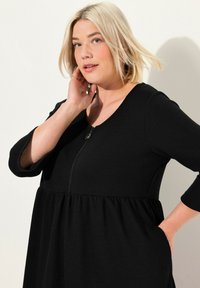Schwarzes Kleid aus weichem Stoff, mit Rundhalsausschnitt, langen Ärmeln, Reißverschluss und ausgestelltem Bund mit Taschen.
