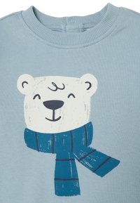 Hellblaues Sweatshirt mit einem bedruckten Eisbären, der einen Schal trägt. Der Bär hat ein weißes Gesicht und einen lächelnden Ausdruck, mit einfachen, fetten Linien.