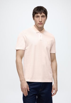 Homme debout devant un fond uni, portant un polo rose clair à manches courtes et un jean foncé, regardant droit devant lui.