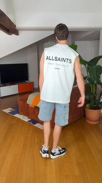 Άνδρας που φοράει λευκό αμάνικο μπλουζάκι AllSaints και ανοιχτόχρωμα τζιν σορτς στέκεται σε σαλόνι με πορτοκαλί καναπέ και μεγάλο φυτευμένο φυτό.