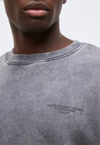 Κοντινό πλάνο ενός ατόμου που φοράει γκρι φούτερ με στρογγυλή λαιμόκοψη και μικρό μαύρο κείμενο τυπωμένο στο στήθος.