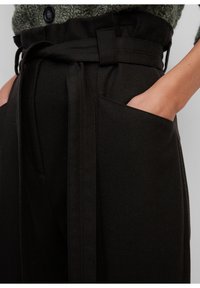 Pantalons noirs taille haute avec une large ceinture, dotés de poches latérales et d'un tissu texturé. Le tissu a un léger éclat et une coupe structurée.