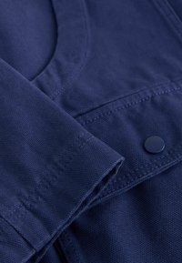 Tamsiai mėlyna audinio medžiaga su tekstūruotu pynimu, su sulenktu rankogaliu ir apvalia iškirpte. Prie siūlės yra tamsus mygtuko akcentas.