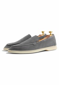 Grijze suede instappers met een rubberen zool, voorzien van een ronde neus en gestikt detail. Houten schoenleesten zichtbaar aan de binnenkant.