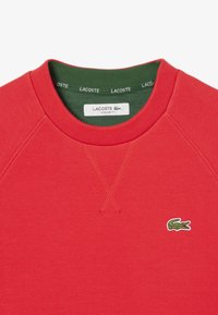 Felpa rossa con finitura testurizzata, colletto verde a costine e logo Lacoste ricamato. Presenta un classico scollo a girocollo e maniche raglan.