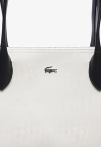 Witte textuurrijke stoffen tote bag met zwarte leren accenten. Heeft een zilveren krokodillenlogo en dubbele handgrepen. Rechthoekige vorm.