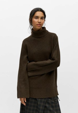 Femme aux cheveux foncés porte un pull à col roulé brun foncé et une jupe à carreaux noire et grise sur un fond clair uni.