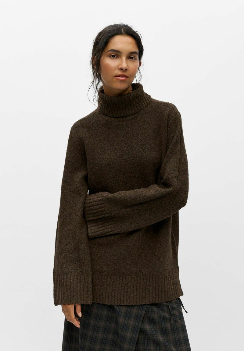 Femme aux cheveux foncés porte un pull à col roulé brun foncé et une jupe à carreaux noire et grise sur un fond clair uni.