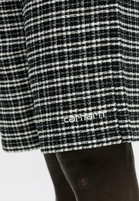 Ύφασμα ασπρόμαυρου καρό με κεντημένο το λογότυπο Carhartt κοντά στο ρέλι πάνω σε σκουρόχρωμο πόδι.