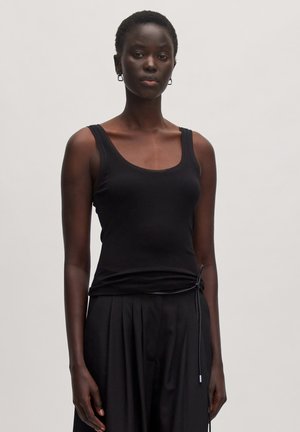Schwarzes geripptes Top mit breiten Trägern, das eine taillierte Silhouette bietet. Kombiniert mit schwarzen, gefalteten Hosen und einem Kordelzugdetail an der Taille.